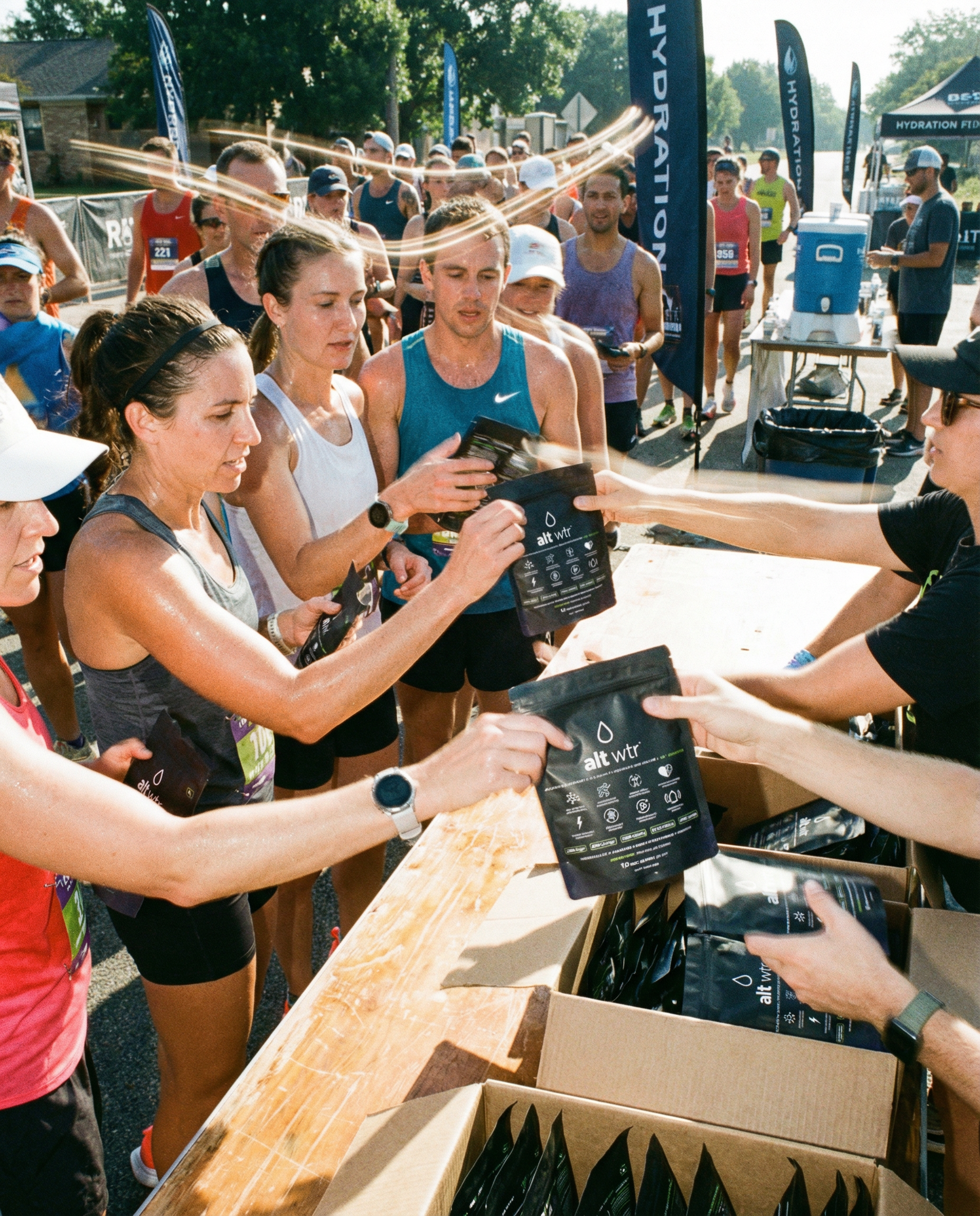 Generated_with_Kive.ai_-_group_of_runners_at_a_running_event_gathering_around_a_rest_station_grabbing_a_bag_of_alt_wtr_being_handed_out_to_the_athletes_participating_in_the_run._day_t.png