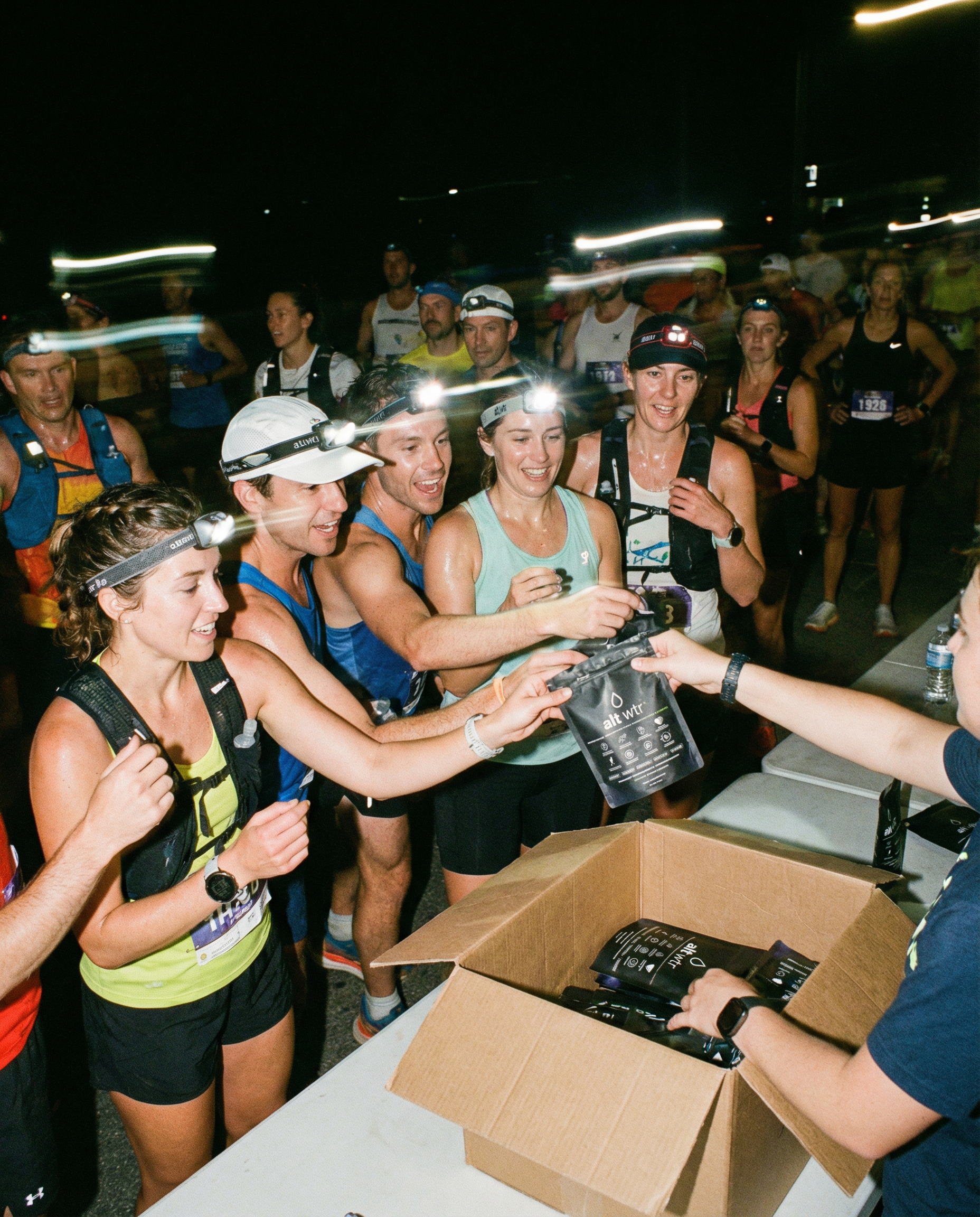 Generated_with_Kive.ai_-_group_of_runners_at_a_running_event_gathering_around_a_rest_station_grabbing_a_bag_of_alt_wtr_being_handed_out_to_the_athletes_participating_in_the_run._night_e4e6d7a2-87dc-4fe3-8009-5d4a945a411f.png